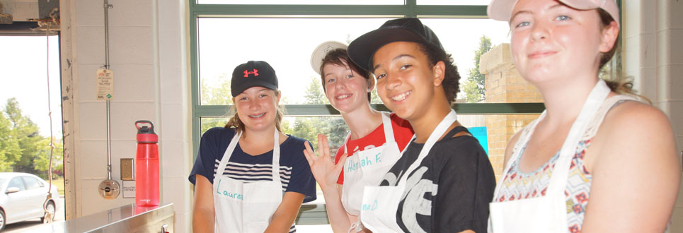 Four female students in a kitchen cooking and wearing hats and white aprons